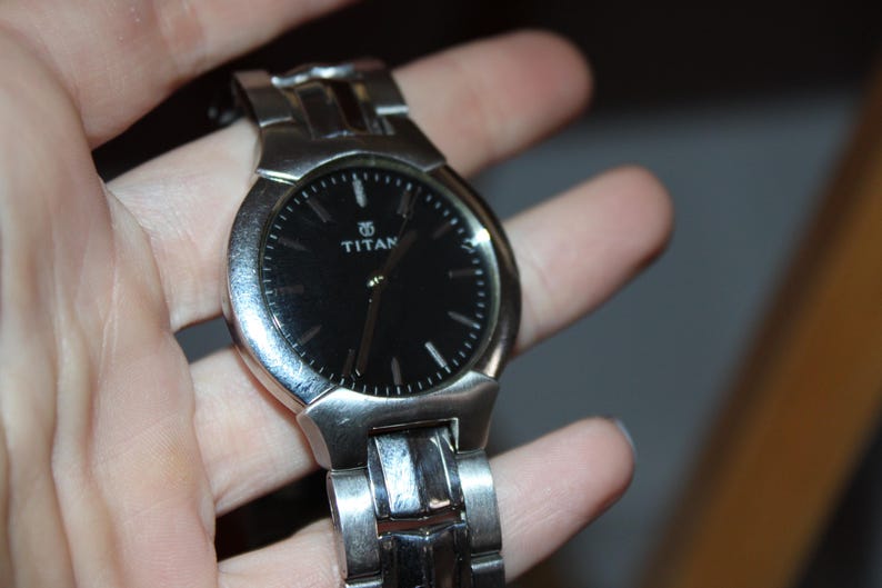May include: A silver-toned Titan wristwatch with a black watch face. The watch has a round face with silver hour markers and hands. The watch band is also silver and appears to be made of metal.