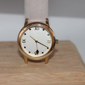 May include: A gold-toned wristwatch with a white face and black Roman numeral hour markers. The watch has gold hands and a light-colored leather band. The watch is displayed on a wooden surface.