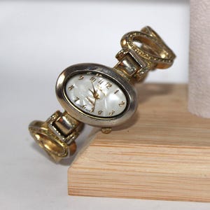 May include: A vintage oval-faced wristwatch with a mother-of-pearl dial. The watch features gold-toned accents on the band and bezel, with a silver-toned face. The watch has a classic design, with the brand name "Salerno" visible on the dial.