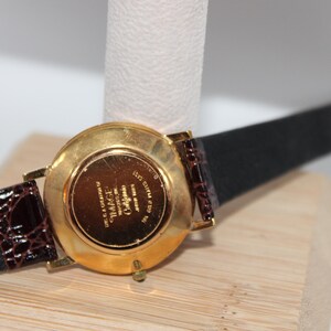 May include: A gold-toned wristwatch with a black leather strap and brown crocodile-patterned leather accents. The watch face is not visible. The watch is displayed on a white cylindrical stand and a wooden surface.