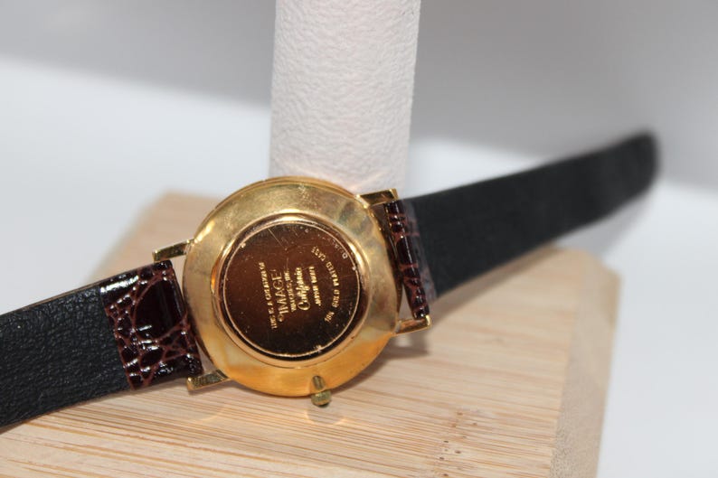 May include: A gold-toned wristwatch with a black leather strap and brown crocodile-patterned leather accents. The watch face is not visible. The watch is displayed on a white cylindrical stand and a wooden surface.