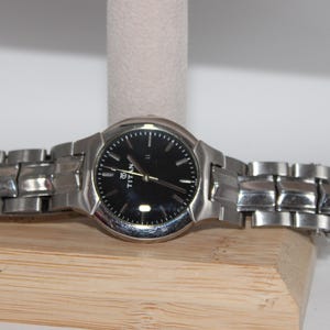 May include: A silver-toned wristwatch with a black watch face. The watch has a metallic band and is displayed on a wooden stand. The watch face has gold-colored hands and markers.