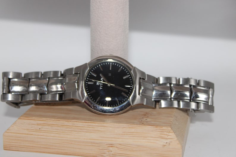 May include: A silver-toned wristwatch with a black watch face. The watch has a metallic band and is displayed on a wooden stand. The watch face has gold-colored hands and markers.