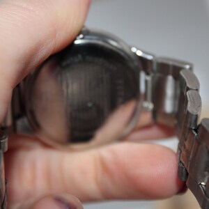 May include: A silver-toned wristwatch with a metal band. The watch face is round and the band is composed of rectangular links. The watch is held in a hand, showing the back of the watch.