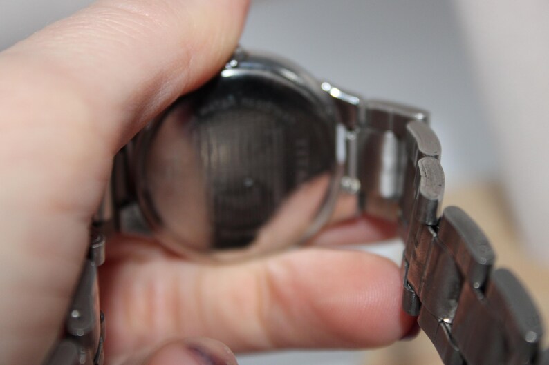 May include: A silver-toned wristwatch with a metal band. The watch face is round and the band is composed of rectangular links. The watch is held in a hand, showing the back of the watch.