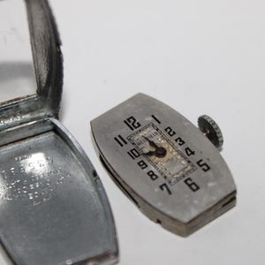 May include: A disassembled vintage silver-toned wristwatch with a rectangular case and a separate watch face. The watch face has black numerals and the brand name "Westfield". The case is open, revealing the watch's interior.