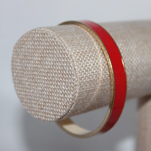 May include: A red and gold bangle bracelet. The bracelet has a bright red enamel band with a thin gold-colored edge. The jewelry is displayed on a beige fabric jewelry stand.