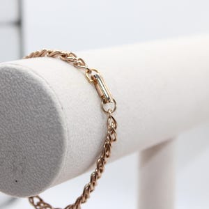 May include: A gold-colored bracelet with a woven chain design. The bracelet is displayed on a white cylindrical jewelry stand. The clasp is a rectangular bar with a gold finish. The bracelet is a delicate piece of jewelry.