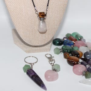 May include: A clear glass vial pendant with a light pink crystal inside, hanging from a black cord. Two keychains with gemstone pendants and a collection of colorful gemstone vials are also visible.