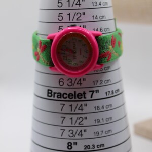 May include: A pink Timex watch with a green and red floral patterned strap, displayed on a bracelet sizer. The watch face is pink with a light-colored dial. The bracelet sizer measures up to 20.3 cm (8 inches).