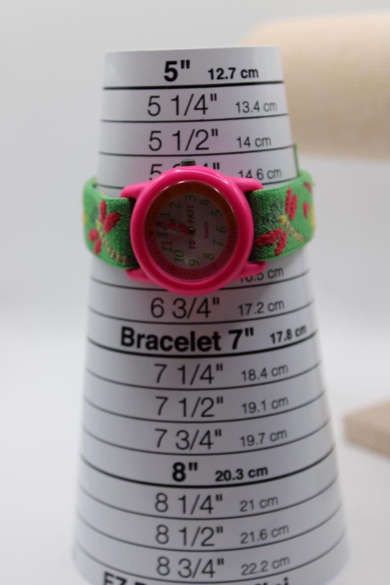 May include: A pink Timex watch with a green and red floral patterned strap, displayed on a bracelet sizer. The watch face is pink with a light-colored dial. The bracelet sizer measures up to 20.3 cm (8 inches).