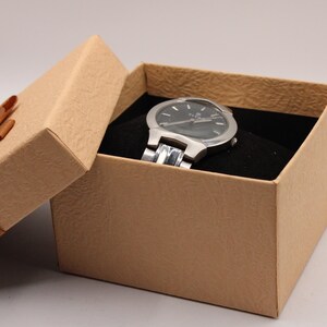 May include: A silver wristwatch with a black watch face rests inside a tan gift box with a brown ribbon. The watch has a silver band and is set against a black velvet interior. The box is slightly ajar.