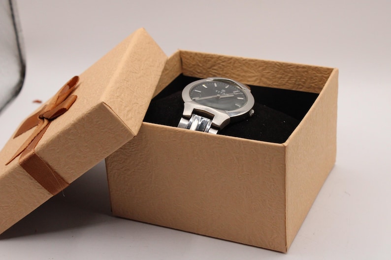 May include: A silver wristwatch with a black watch face rests inside a tan gift box with a brown ribbon. The watch has a silver band and is set against a black velvet interior. The box is slightly ajar.