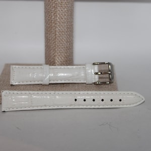 May include: Two-piece white watch band with a textured, crocodile-like pattern. The band features a silver-colored buckle and several holes for adjustment. The watch band is displayed on a neutral-colored stand.