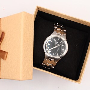 May include: A silver and brown wristwatch with a black face rests inside an open, tan gift box with a brown ribbon and bow. The watch has a metallic band and a dark dial with silver accents.