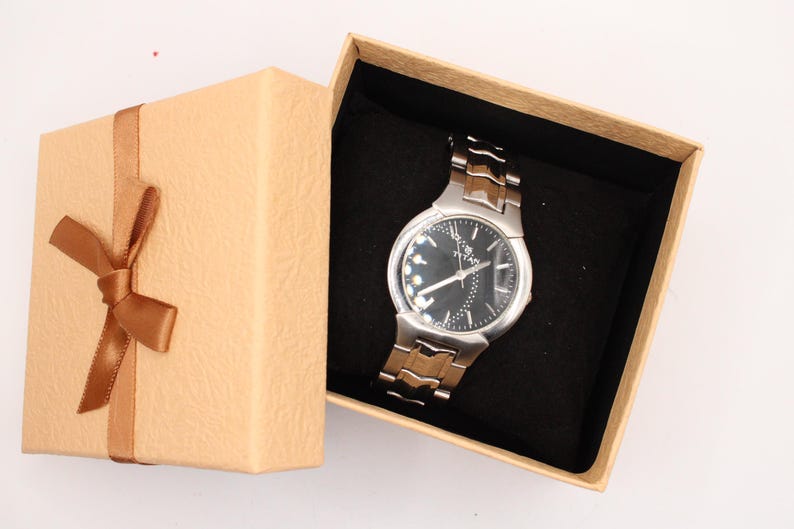 May include: A silver and brown wristwatch with a black face rests inside an open, tan gift box with a brown ribbon and bow. The watch has a metallic band and a dark dial with silver accents.