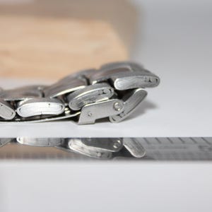 May include: Close-up of a silver metal watch band with a clasp. The band is composed of multiple rectangular links. The watch band is on a white surface with a ruler in the foreground and a wooden block in the background.