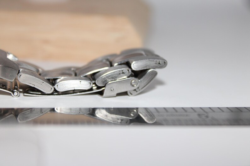 May include: Close-up of a silver metal watch band with a clasp. The band is composed of multiple rectangular links. The watch band is on a white surface with a ruler in the foreground and a wooden block in the background.