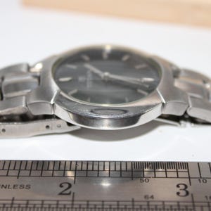 May include: A silver-toned wristwatch with a dark gray face and a stainless steel band. The watch is positioned next to a stainless steel ruler, showing measurements in inches. The watch's face has silver hands and markers.
