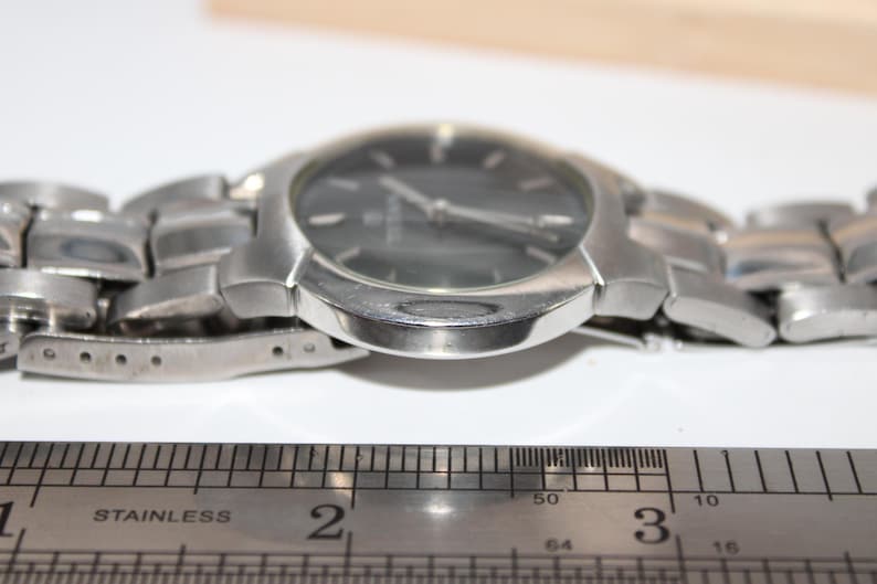 May include: A silver-toned wristwatch with a dark gray face and a stainless steel band. The watch is positioned next to a stainless steel ruler, showing measurements in inches. The watch's face has silver hands and markers.