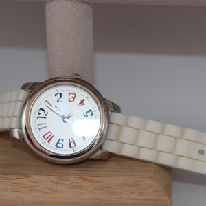 May include: A silver wristwatch with a white face and a white band. The watch face has colorful numbers and hands. The watch is resting on a wooden surface.