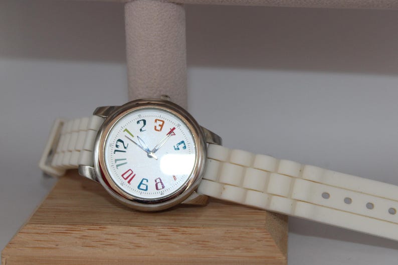 May include: A silver wristwatch with a white face and a white band. The watch face has colorful numbers and hands. The watch is resting on a wooden surface.