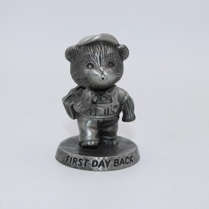 May include: A pewter figurine of a bear cub wearing overalls and a cap, holding a book. The figurine stands on a round base with the words "FIRST DAY BACK" engraved on it. The bear has a surprised expression.