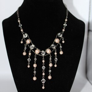 May include: A silver-toned necklace with a delicate design. The necklace features alternating clear crystals and pale pink pearls. Dangles of pearls and crystals hang from the main chain, creating a layered effect. The necklace is displayed on a black mannequin.