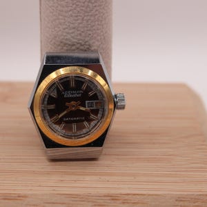 May include: A vintage Accetron Electra watch with a black face and gold-tone bezel. The watch has a silver-tone case and a date display. The watch is displayed on a wooden surface.