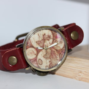 May include: A wristwatch with a burgundy leather band and a round face. The watch face features a map design with a brass-colored bezel. The band has brass-colored rivets and a buckle. The watch is resting on a wooden surface.