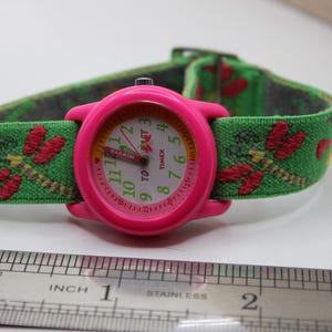 May include: A pink Timex watch with a green strap decorated with red dragonfly patterns. The watch face has white numerals and hands, with the word "TIMEX" visible. The strap is about 2.5 cm wide.
