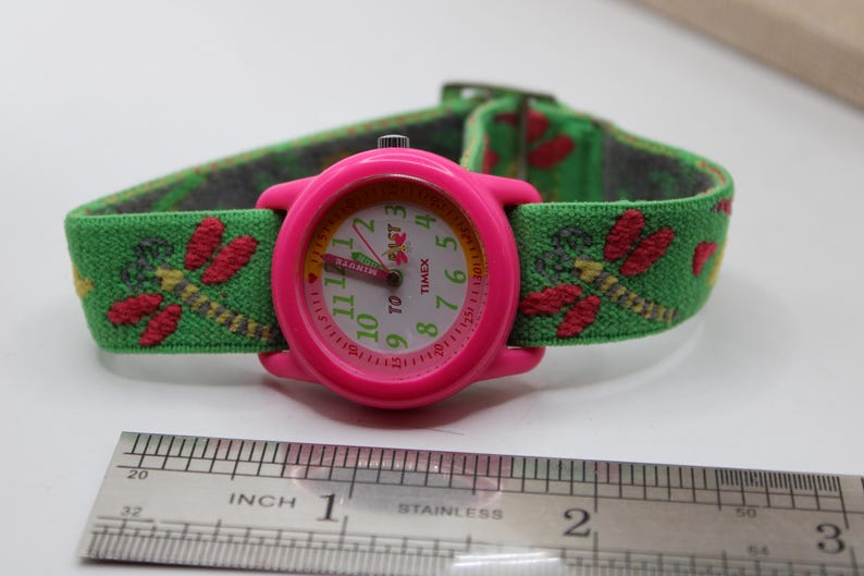 May include: A pink Timex watch with a green strap decorated with red dragonfly patterns. The watch face has white numerals and hands, with the word "TIMEX" visible. The strap is about 2.5 cm wide.