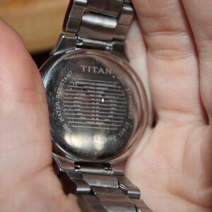May include: A silver-toned Titan wristwatch with a stainless steel band. The watch back is visible, displaying the brand name and the words "Water Resistant". The watch is held in a hand.