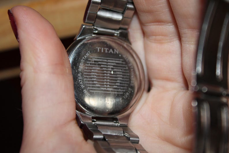 May include: A silver-toned Titan wristwatch with a stainless steel band. The watch back is visible, displaying the brand name and the words "Water Resistant". The watch is held in a hand.