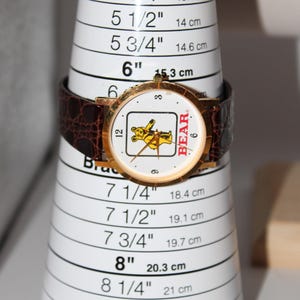May include: A gold-toned wristwatch with a brown leather band is displayed on a white bracelet sizing cone. The watch face features a cartoon bear graphic and the word "BEAR". The cone measures up to 8 3/4 inches.