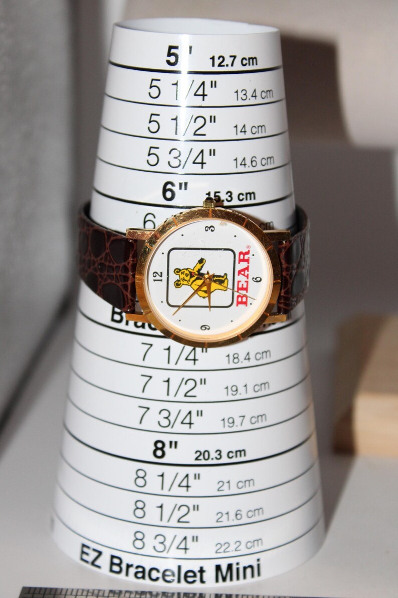 May include: A gold-toned wristwatch with a brown leather band is displayed on a white bracelet sizing cone. The watch face features a cartoon bear graphic and the word "BEAR". The cone measures up to 8 3/4 inches.