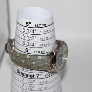 May include: A silver wristwatch with a patterned band is displayed on a white bracelet sizer. The sizer shows measurements in inches and centimeters, with the watch resting at the 6-inch (15.3 cm) mark.