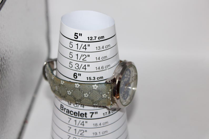 May include: A silver wristwatch with a patterned band is displayed on a white bracelet sizer. The sizer shows measurements in inches and centimeters, with the watch resting at the 6-inch (15.3 cm) mark.