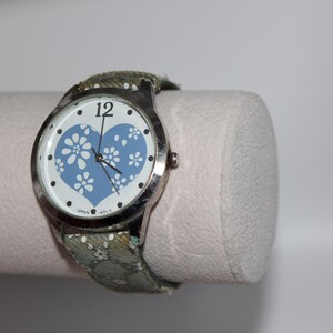 May include: A silver wristwatch with a white face featuring a blue heart design filled with white floral patterns. The watch has a green floral patterned band. The watch is displayed on a white cylindrical stand.