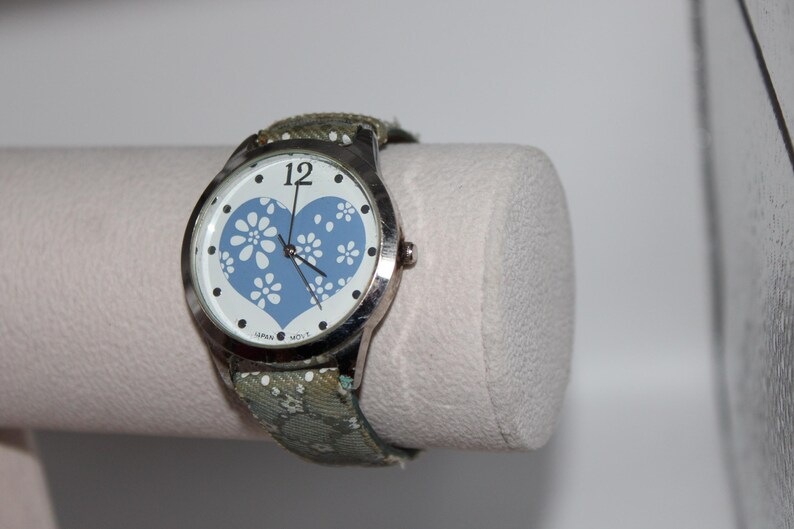 May include: A silver wristwatch with a white face featuring a blue heart design filled with white floral patterns. The watch has a green floral patterned band. The watch is displayed on a white cylindrical stand.
