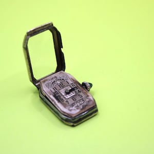 Puede incluir: Un reloj de pulsera antiguo plateado con una esfera rectangular y una tapa con bisagras. La esfera del reloj muestra números y manecillas, con una pequeña corona en el lateral. El reloj está sobre un fondo verde brillante.