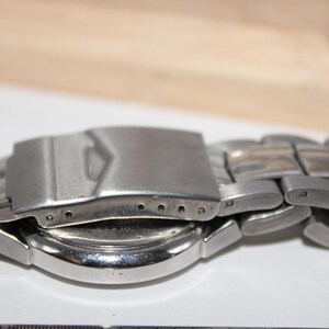 May include: Close-up of a silver-toned wristwatch. The watch band is made of interlocking metal links. The clasp is open, revealing the watch face and the back of the watch. The background is a light color.