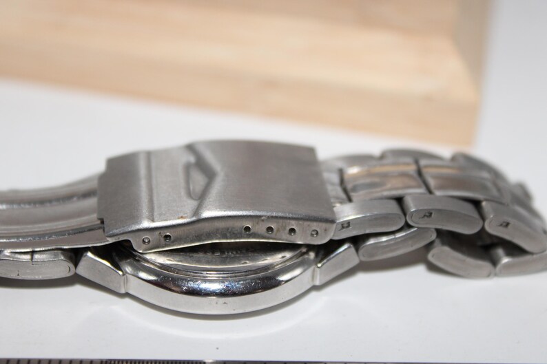 May include: Close-up of a silver-toned wristwatch. The watch band is made of interlocking metal links. The clasp is open, revealing the watch face and the back of the watch. The background is a light color.