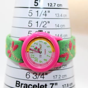 May include: A pink Timex watch with a green fabric strap with a floral design. The watch face is white with black numerals and the words "TO PAST" and "TIMEX". The watch is on a white measuring tool with measurements in cm.