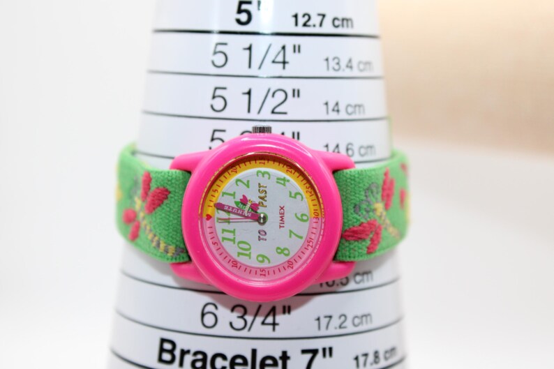 May include: A pink Timex watch with a green fabric strap with a floral design. The watch face is white with black numerals and the words "TO PAST" and "TIMEX". The watch is on a white measuring tool with measurements in cm.