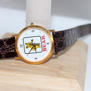 May include: A gold-toned wristwatch with a white face featuring a yellow bear graphic and the word "BEAR." The watch has black hands, a brown leather band, and the numbers 3, 6, and 12. The band has a crocodile pattern.