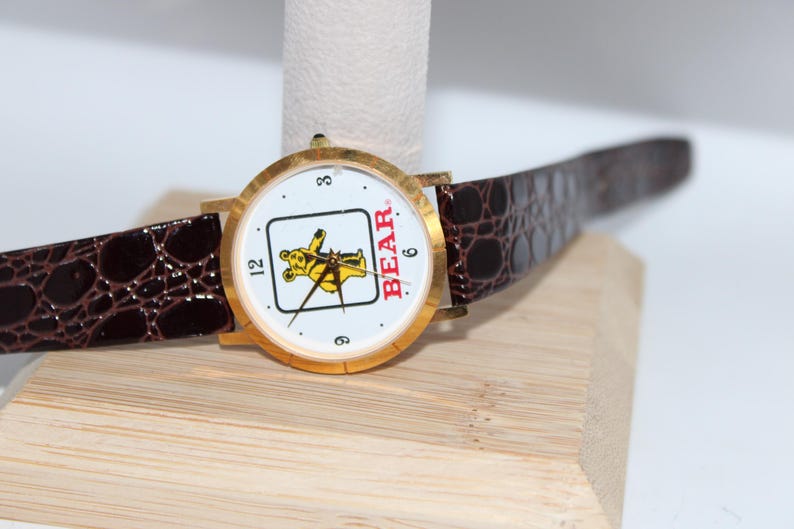May include: A gold-toned wristwatch with a white face featuring a yellow bear graphic and the word "BEAR." The watch has black hands, a brown leather band, and the numbers 3, 6, and 12. The band has a crocodile pattern.