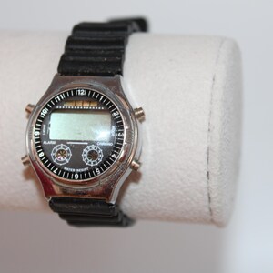 May include: A digital wristwatch with a black rubber band. The watch face is black with silver accents and a digital display. The watch has buttons for alarm and chrono functions. The watch is water resistant.