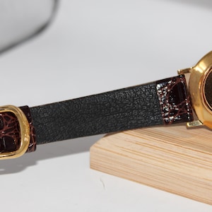 May include: A gold-toned watch with a brown leather band. The watch band has a black textured center and brown crocodile-style accents near the buckle. The watch face is gold and has the text "IMAGE California Japan 1991".