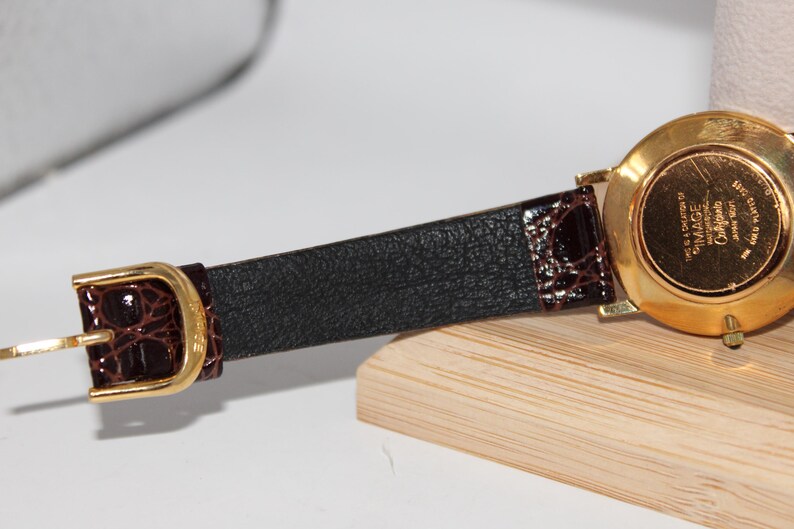 May include: A gold-toned watch with a brown leather band. The watch band has a black textured center and brown crocodile-style accents near the buckle. The watch face is gold and has the text "IMAGE California Japan 1991".